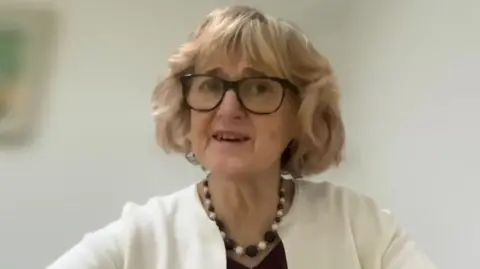 A woman with a short blonde bob wearing black rimmed glasses and a cream cardigan sat in front of a camera.