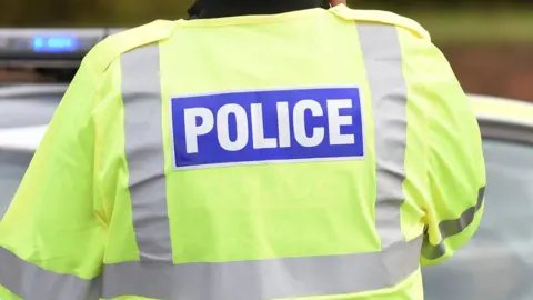 Officer in fluorescent police jacket with blue lights on a police car in the background.