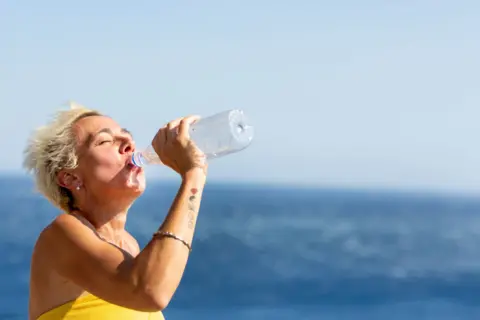 A woman with short blonde hair drinks from a plastic water bottle by the sea.