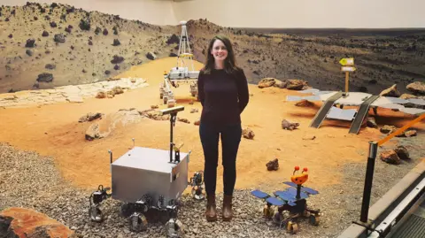 A woman wearing a brown jumper and black trousers stands on a simulated surface of Mars with orange sand and stones, with prototypes at her feet