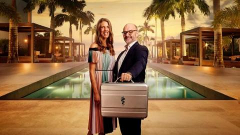 Couple stood in front of pool holding suitcase as part of ITV show Fortune Hotel