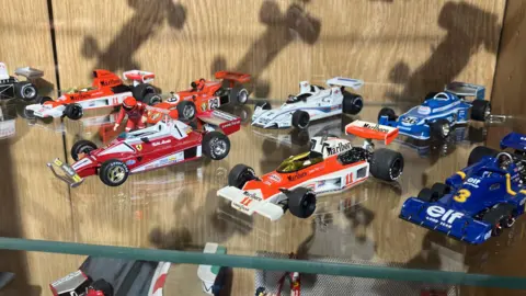 BBC/Jack Hadaway-Weller A glass display shelf holds detailed miniature Formula One race cars arranged neatly in rows.
The models feature bright liveries in red, white, blue, and orange, with visible sponsor logos and numbers.
Reflections on the glass and shadows on the wooden backing add depth to the carefully curated collection.
