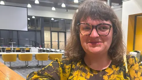 A woman stares at the camera. She wears a black and yellow top. She has big rimmed glasses and a lip piercing. In the background are officials wearing yellow lanyards and a number of desks. 