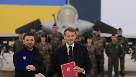 Reuters Ukrainian President Volodymyr Zelensky (left) and French President Emmanuel Macron (right) shake hands after signing a letter of intent at the Villacoublay air base near Paris. French soldiers and a war plane are seen in the background.