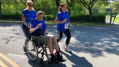 Tom Davies sitting in a wheelchair being pushed by a woman with blonde hair and wearing sun glasses, while his mother, with brown hair, runs alongside them. All three people are wearing blue t-shirts to signal they are participating in the race.