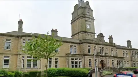 A long large Victorian building takes up most of the image with steps leading up to the entrance and a ramp to one side. A large clock tower sits in the centre of the building and there are several chimneys