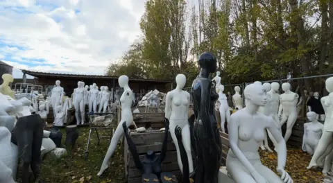 An array of mannequins are standing together outside. Some are glossy others are matte. Some are perched on wooden slats whilst others are sat on boxes.