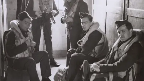 IWM Duxford A black and white photo showing five pilots in a locker room at RAF Duxford in World War Two, getting ready for a mission. They are wearing uniform, and life guards. One man is sitting on the left and two on the right, each wearing a cap. They are looking towards the camera. Behind them are other standing pilots. 