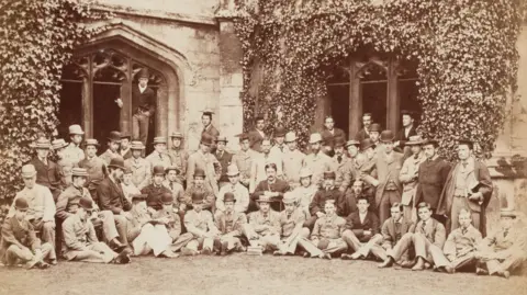 Dominic Winter Auctioneers A black and white group photo taken in the cloisters of Magdalen College, Oxford, in 1876. The men in the image are all well dressed.