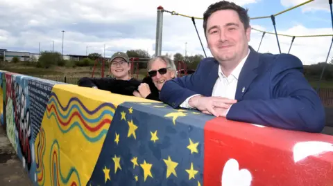 Open Door Art Jess Taylor has a brown cap, black top and glasses and is smiling. Councillor Graham Wilson has white hair, black glasses and a black top. He is smiling. Mayor Chris Cooke has brown hair, a white shirt and a formal blue jacket. He is smiling. They are leaning on multi coloured wall.