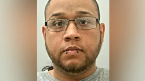 Robert Court's mugshot. He has dark hair and a short beard, black framed wire glasses and what looks like two big stitches through his bottom lip