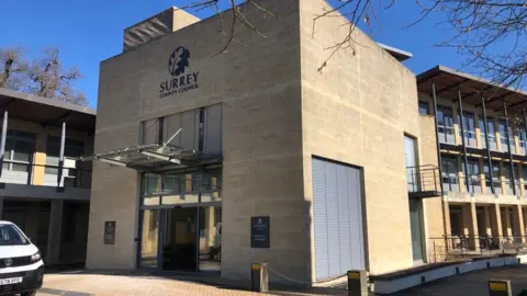 Surrey County Council headquarters at Woodhatch Place in Reigate. Building has a square shape with sand coloured bricks. The entrance door is glass. The words 'Surrey County Council' and a logo can be seen. A white vehicle is to the left of the image.