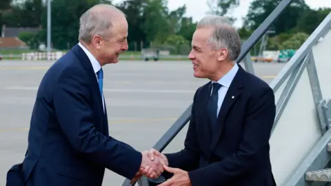 PA Media Canadian Prime Minister Mark Carney is welcomed by Taoiseach Micheal Martin at Dublin airport at the start of his visit to Ireland. They are shaking hands atg the bottom of plane steps.