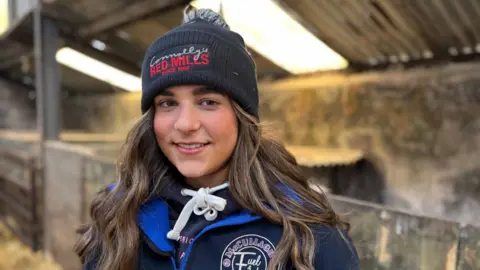 A young woman stands in an animal shed. She is warmly dressed overalls over a hooded top. She is wearing a woolly hat and has long dark hair underneath. She is smiling.