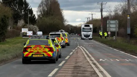 BBC/Crispin Rolfe A row of police vehicles along the A1079 at Wilberfoss, with officers investigating at the scene. A white HGV is stationary on one side of the road. Two police officers with equipment in their hands are by its side.