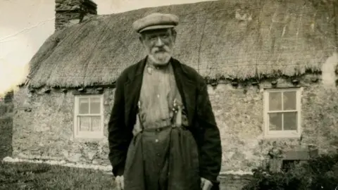 MNH An old photo of Harry Kelly, a man in a baggy blazer, and tweed farmin cap, he stands in front of the thached cottage, now known as Harry Kelly's cottage.