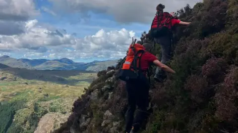 KMRT Teddy in the rucksack of a rescuer who is coming down a mountain with another person. There is a dramatic view of the Lake District fells behind them.