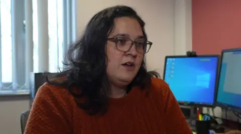 Dr Rachel Ali, who has long brown hair and is wearing glasses and a burnt orange jumper, is sitting at a desk in a consulting room.