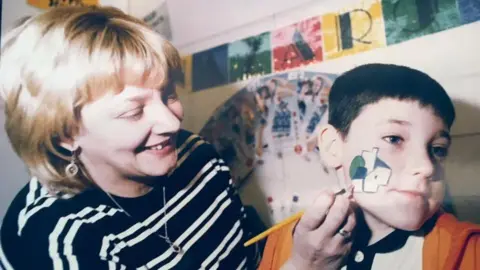 Handout A young Lauren Staton painting an abstract design on a child's face. Lauren has short blonde hair and is wearing a black and white long-sleeved striped top. The child has short dark hair and has his face turned to the side.