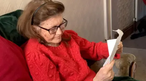 A woman with brown shoulder length hair sits on a green sofa in a living room and looks at a letter. She is wearing a red knitted jumper and glasses. 