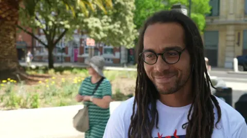 A man in a white shirt with dreads smiles whilst standing on street outdoors.