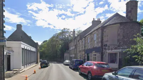 Union Street in Wick with cars parked on both sides of the road and some police no-waiting cones. There are brick houses down the right hand side and what looks like some kind of showroom on the left with trees in the distance.
