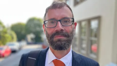 Deputy Aidan Matthews - A man with black and grey hair and a big bushy grey and black beard. he is wearing wonky spectacles and a blue pinstriped suit on top of a white shirt and orange tie. 