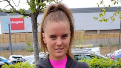 Family handout Holly Newton standing in a car park. She has long brown hair tied up in a ponytail and is smiling at the camera.