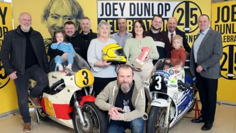 Pacemaker Two motorbikes, one white and red, and the other blue and white sit next to each other in front of a yellow Joey Dunlop 25 years board with his face printed on it. There are ten people (8 men and 2 women) standing around the bikes dressed in a range of clothing from black, grey, blue, green and beige. There are also two small children sitting on the bikes, a girl with brown hair wearing a blue tracksuit and a boy with an orange jumper.
