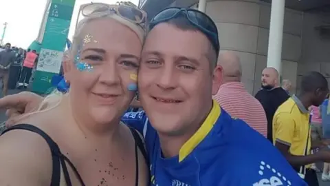 Emma Miller and James Miller are outside Wembley Stadium. James is wearing the Warrington Wolves jersey and Emma is wearing a black strappy top and has sparkly face paint in the team's colours of yellow and blue. A number of other people can be seen in the background.