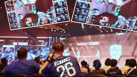 Getty Images Spectators are shown in an arena looking at teams competing during the 2026 Esports World Cup in Paris. Four large screens can be seen showing the action taking place. 