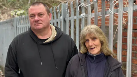 Simon Thake/BBC A man in a dark fleece stands next to an elderly woman with long greying hair. Behind them is a spiky metal fence.