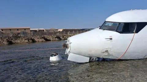Starsky Aviation A damaged aircraft rests partly submerged on a shallow shoreline, with waves washing against its wing as it lies in clear, shallow water under a bright blue sky.