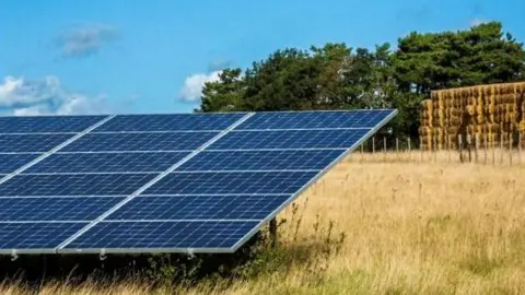 South Kesteven District Council A row of solar panels in a field with a stack of bales and trees in the background.