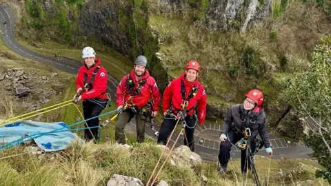 Avon and Somerset Search and Rescue Four people are on the edge of Cheddar Gorge, fastened to ropes. They are wearing red jackets and red helmets.