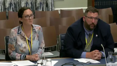 NI Assembly Sara has dark brown hair. She is wearing a blue patterned shirt with pink flowers. Michael is sitting beside her, he has brown hair and is wearing a navy suit, blue shirt and green tie.
