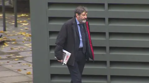 BBC Alan Harris wearing a black coat with a red inner lining, dark suit, blue shirt and a dark blue tie with yellow markings on it walks past a green metal structure which has ventilation slots on the exterior. He is carrying a couple of notepads. Mr Harris has a greying beard and greying hair.