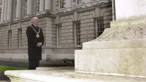 Pacemaker Alex Maskey stands in front of the Cenotaph outside Belfast City Hall in  July 2002.  He is wearing a long black coat, a dark suit and his gold lord mayor's chain. 