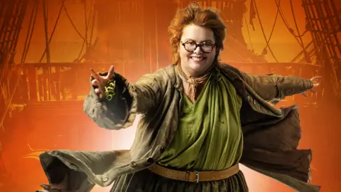Actress Jayde Adams in full costume in a promotional picture for the show Treasure Island at Bristol Old Vic. She is wearing a large brown jacket and green shirt and behind her is a glowing orange scene and the silhouette of a pirate ship