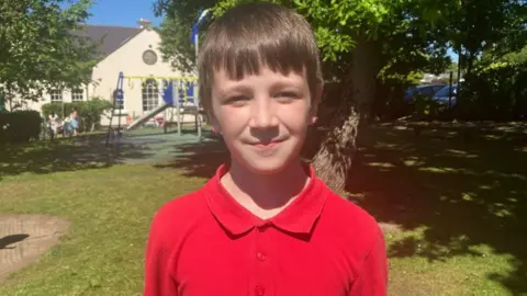 Harry is wearing a bright red polo top and has short brown hair. He is looking at the camera. He is standing on grass with a tree in the background, further in the distance the school building can be seen.