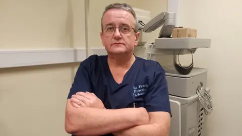 Man sitting with his arms folded looking at the camera. He is wearing navy medical scrubs