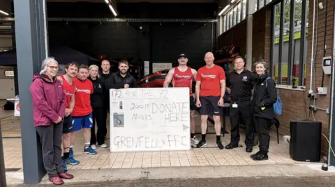 Matt Broadbent Seven men and three women standing in the entrance to the fire station. They have a large sign in front of them which reads "72 for the 72.. Donate here. Grenfell FFC"