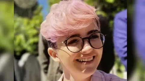 Imogen Nunn is smiling and looking off camera. She has pale pink hair and a pink checked jacket on, and is wearing dark rimmed round glasses.
