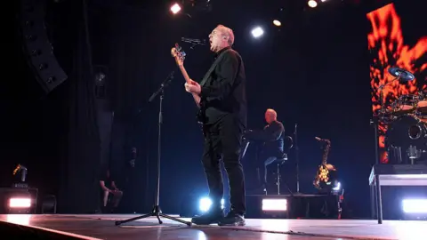 Getty A man dressed in all black sings to a microphone on a stage as he plays an electric guitar. Behind him is another man who is sitting on a chair next to a saxophone.