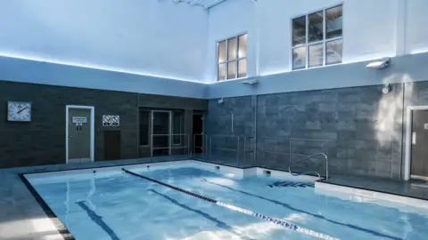 David Lloyd An indoor swimming pool. It has blue tiles and blue and white lane dividers. The walls are tiled grey.