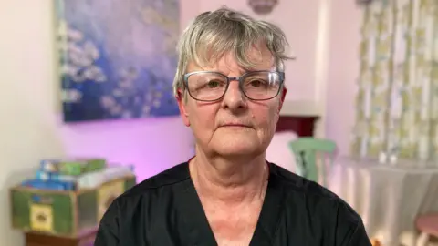 Jill has short wavy grey hair and wears silver-rimmed glasses. She is looking straight at the camera and wears black scrubs. She is sitting in a communal room at the care home, with a painting, a box of jigsaws and a table and chairs behind her.