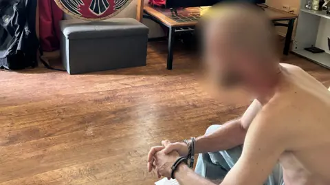 A shirtless man with a blurred face wearing handcuffs sits in a living room
