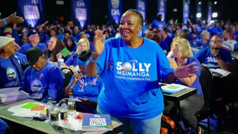 Gallo via Getty Images A woman wearing a blue T-shirt that says 'vote Solly Msimanga' claps her hands and smiles. A sea of delegates sitting at tables can be seen behind her.