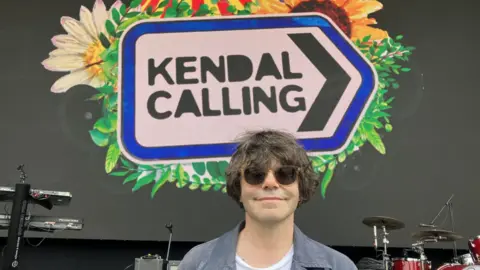Tim Burgess, wearing sunglasses and a blue coat, smiles underneath a Kendall Calling festival sign