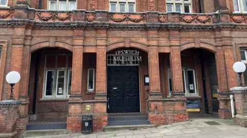 The entrance into Ipswich Museum. It is a red brick building with a large black door. The name of the museum hangs over the door.